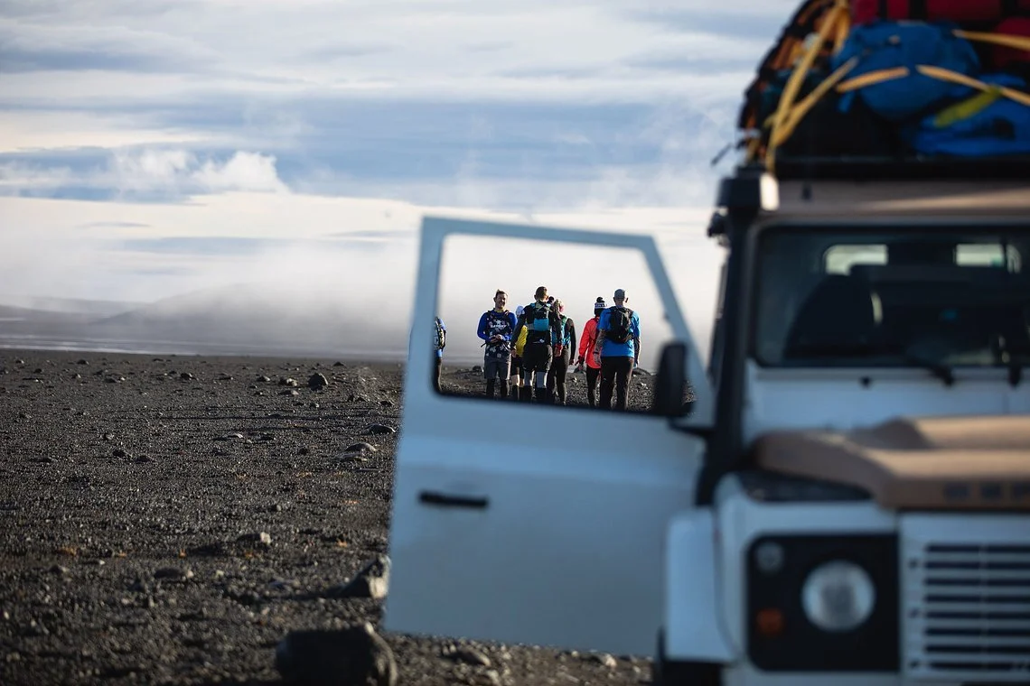 Group of hikers with backpacks walking across volcanic terrain, seen through an open 4x4 vehicle door