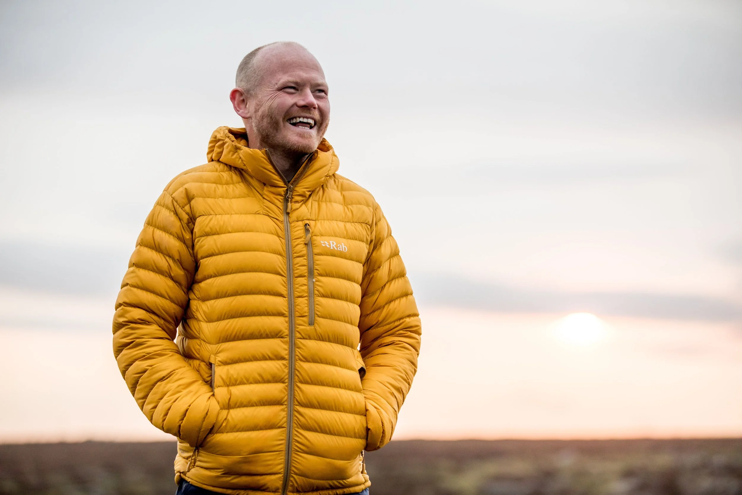 Danny Bent smiling outdoors, wearing a yellow jacket, founder and speaker who brings people together through movement and shared challenge