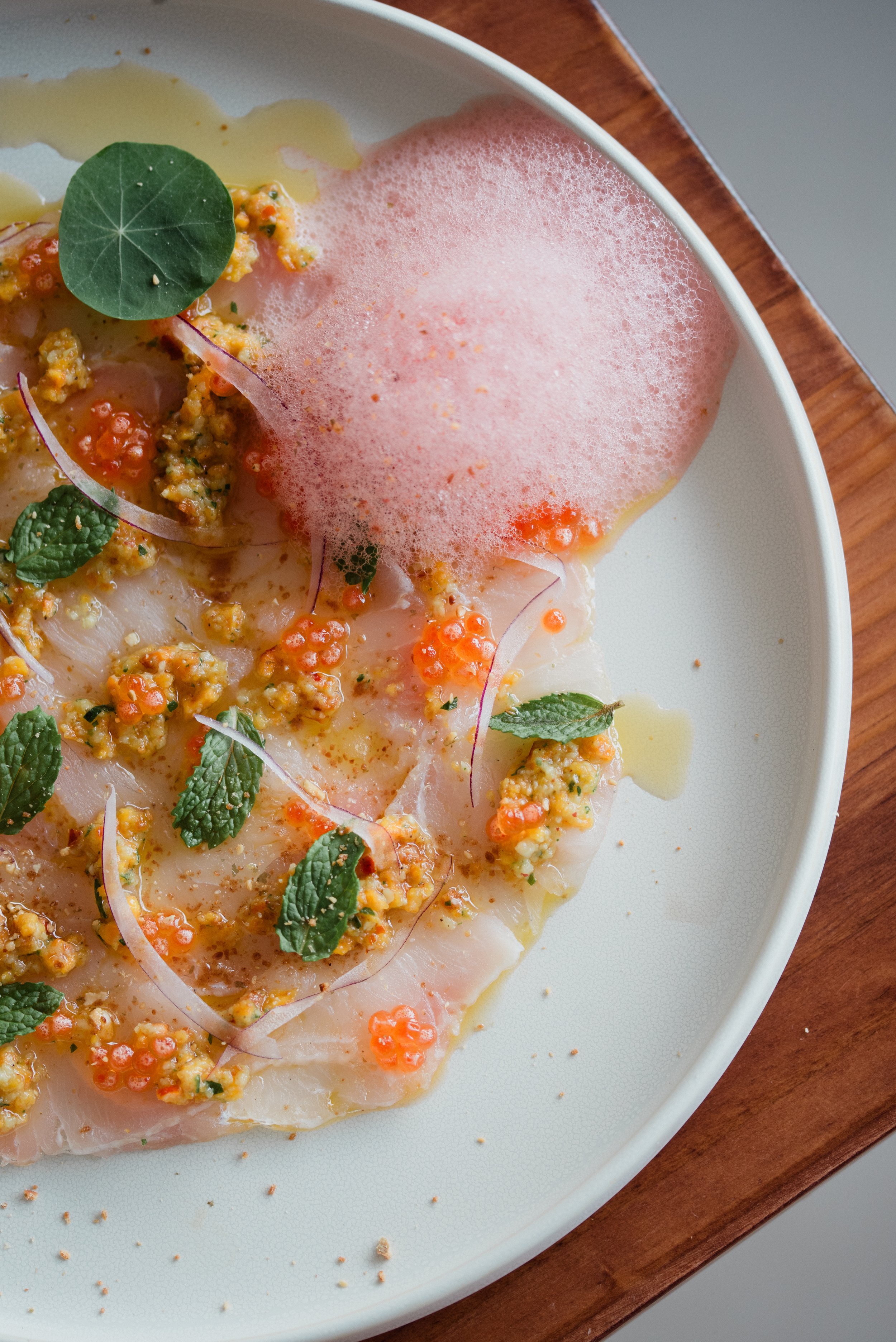 A dish of raw fish with garnishes, topped with a pink foam, microgreens, and small orange roe, served on a white plate on a wooden table.