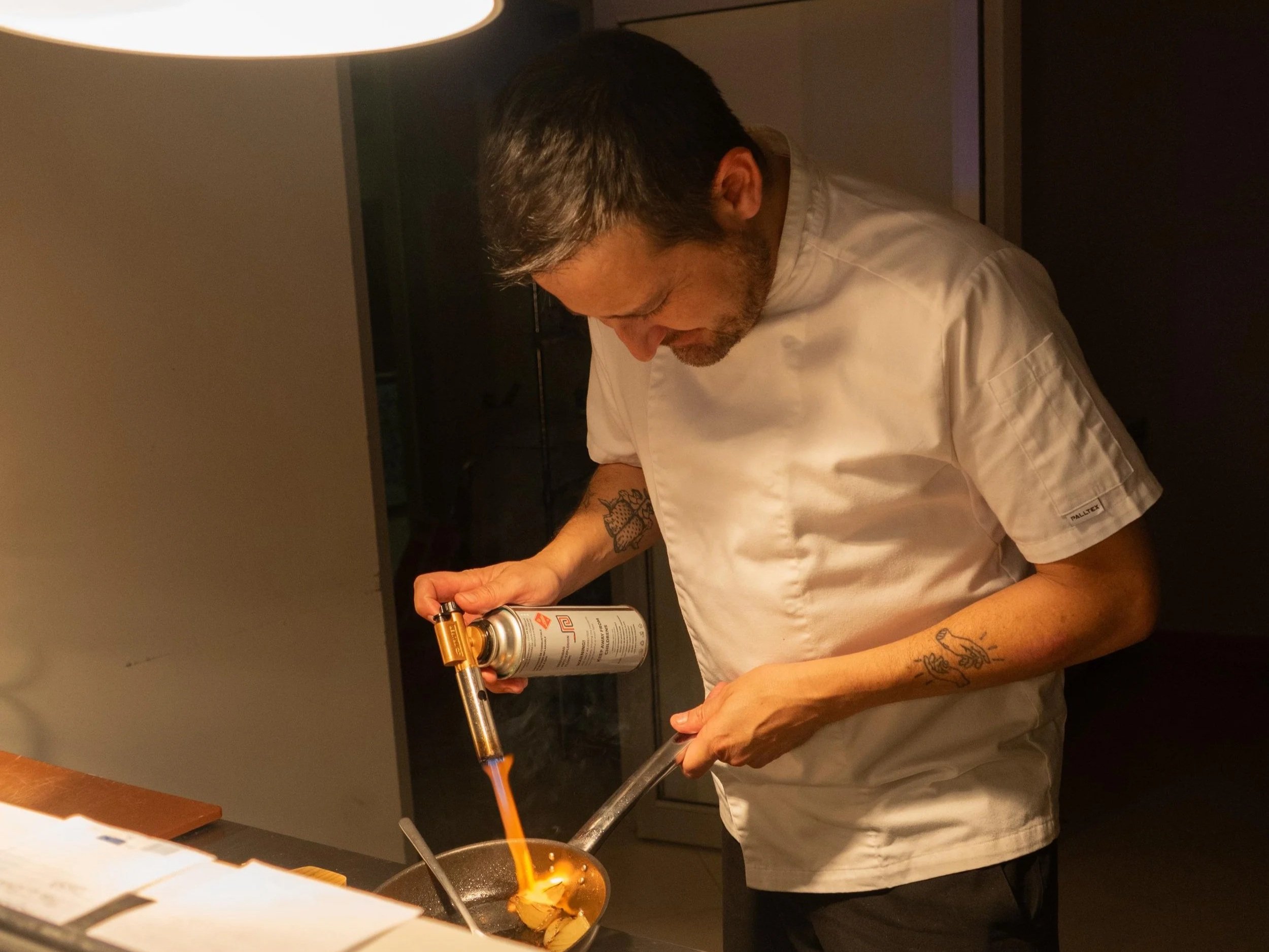 A chef in a white chef's coat using a butane torch to caramelize sugar in a kitchen.
