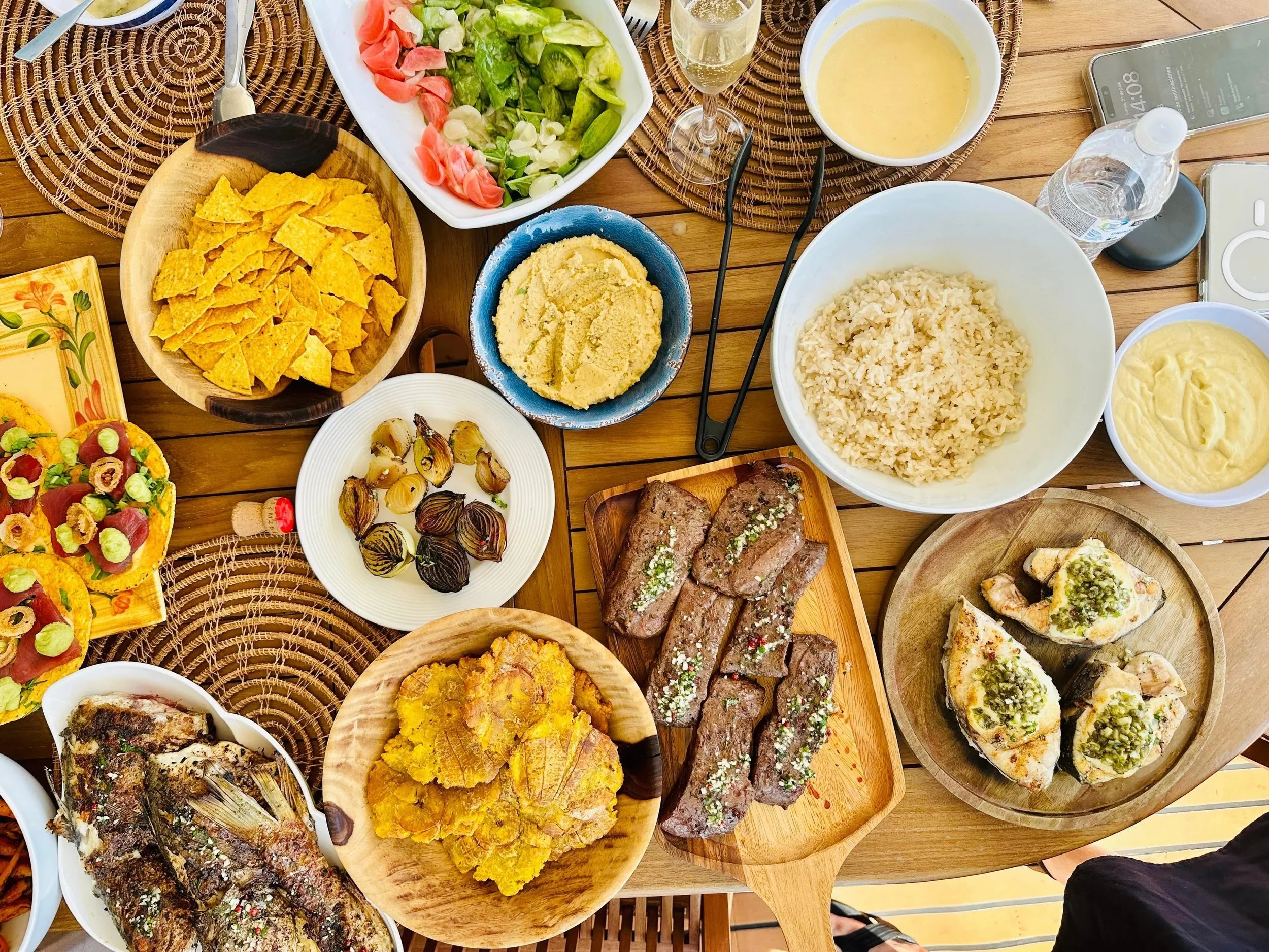 A table filled with various Mexican dishes, including grilled fish, tacos, quesadillas, grilled vegetables, rice, guacamole, salsa, chips, sliced onions, deep-fried tofu, and drinks like champagne and water.