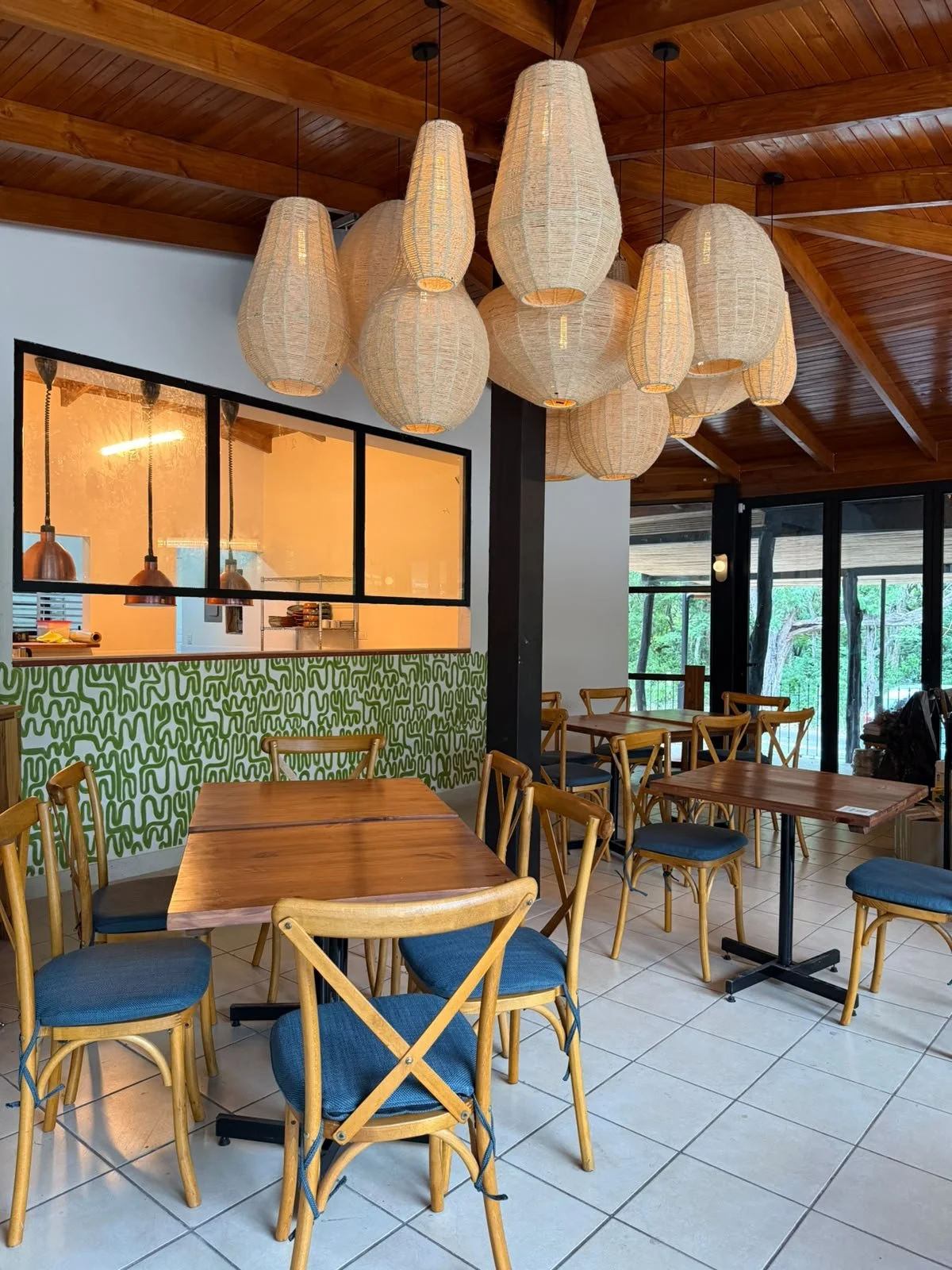 Interior of a modern restaurant with wooden dining tables and chairs with blue cushions. Several large, hanging paper lantern light fixtures are on the ceiling. A green and white abstract patterned wall decorates part of the wall, and a large window with a view of greenery is in the background.
