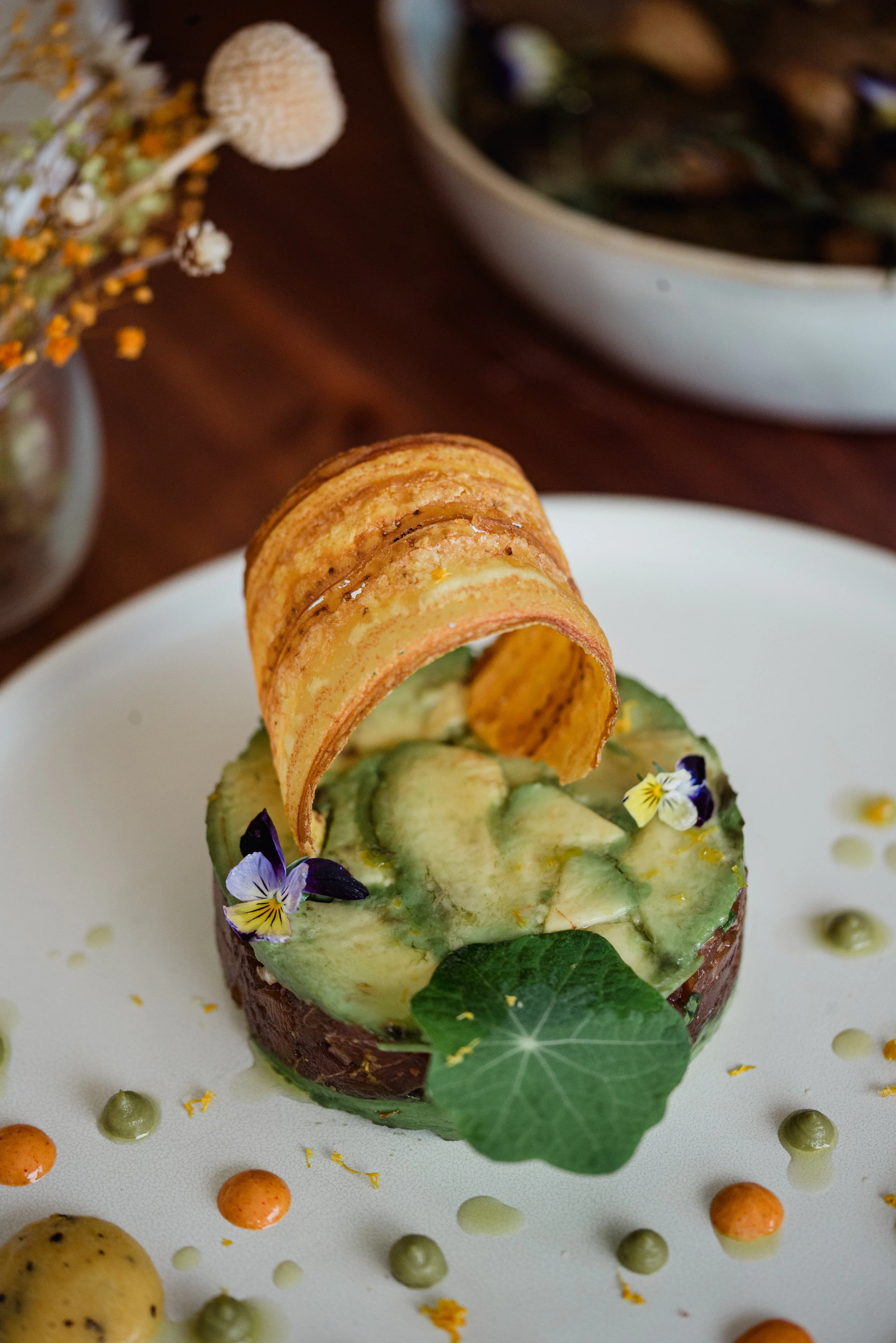 An intricately plated gourmet dish featuring a sliced avocado and a crispy chip arching over it, garnished with edible flowers and a large leaf, on a white plate decorated with small colorful dots of sauces.