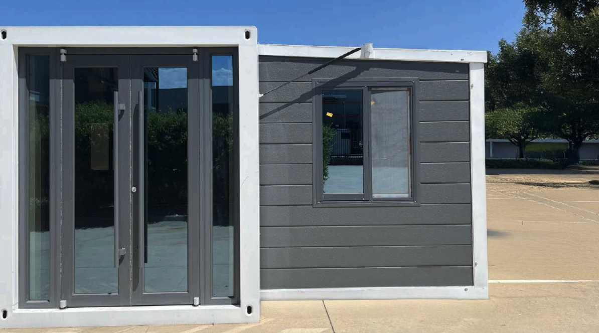 A small modern building with gray siding and large glass door on the left, and a small window on the right, located outdoors on a concrete surface with trees and a parking lot in the background.