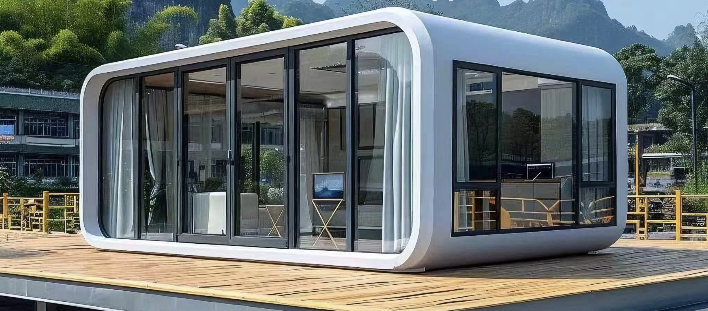 A modern glass house with curved edges on a wooden deck, surrounded by trees and mountains.