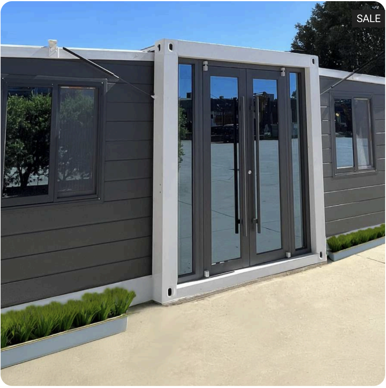 A modern building with dark gray horizontal siding, a glass door with black frame, and windows. There are green plants in a long, white planter along the concrete ground. The sky is clear and blue.