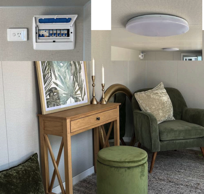 Living room with a wooden console table, a framed tropical leaf art piece, two tall gold candlesticks with white candles, a green upholstered armchair, a matching green round ottoman, and a ceiling light. An electrical panel is exposed on the wall.