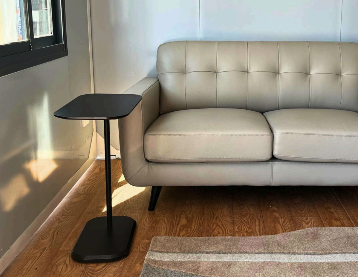 A beige sofa with a tufted backrest next to a small black side table, in a room with wooden flooring and a window on the left.