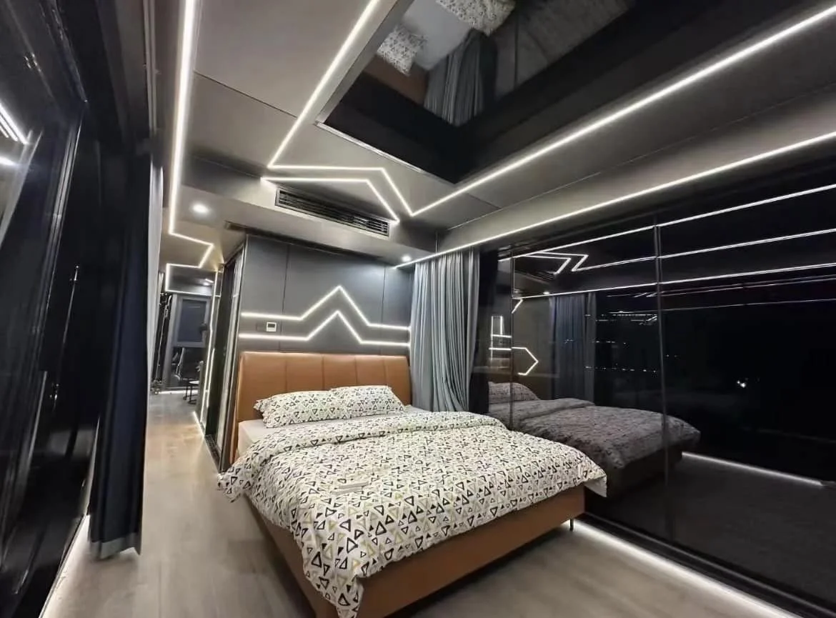 Modern bedroom with a bed that has a brown headboard, geometric patterned bedding, illuminated walls with angular LED strip lights, mirrored closet doors, and floor-to-ceiling windows with curtains.
