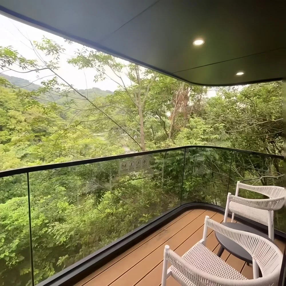A balcony with glass railings overlooking lush green trees and a mountain in the background, with three white chairs and a round side table on a wood deck ceiling with recessed lighting.