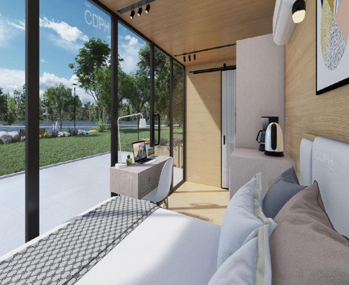 Modern hotel room with large floor-to-ceiling windows showing outdoor greenery, a bed with white linens, a small desk with a TV, and a coffee station in the corner.