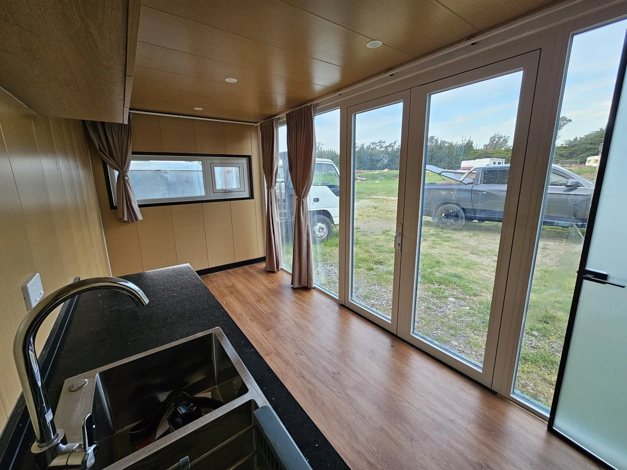 Interior view of a room with large glass doors and windows, showing an outdoor scene with vehicles and grassy area, with wood panel walls and ceiling.