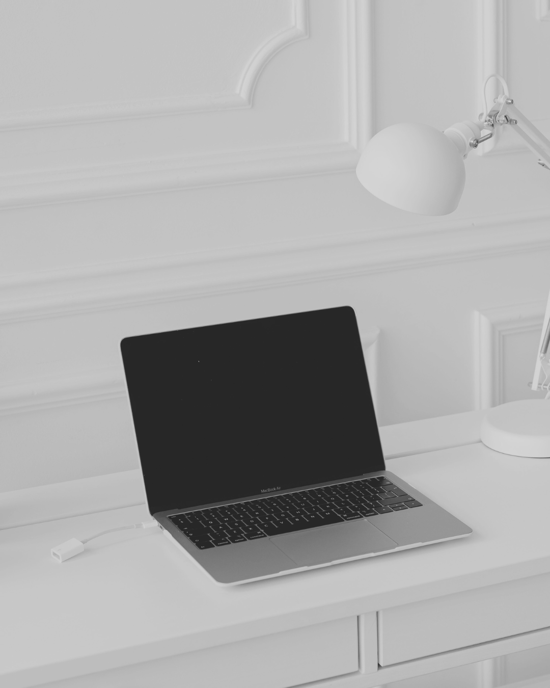 A silver MacBook Air laptop on a white desk with a black screen, connected to a white power cable, under a white wall-mounted desk lamp in a minimalist white room.