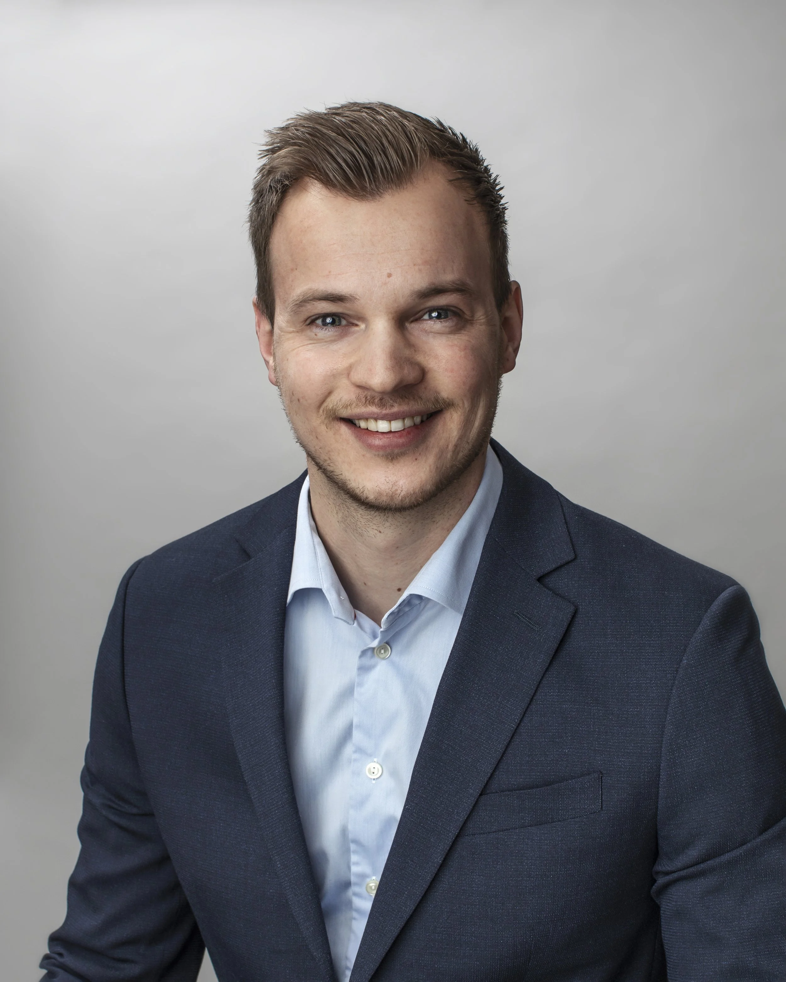 A young man with light brown hair, blue eyes, and light stubble, wearing a dark suit jacket and a light blue shirt, smiling against a light gray background.
