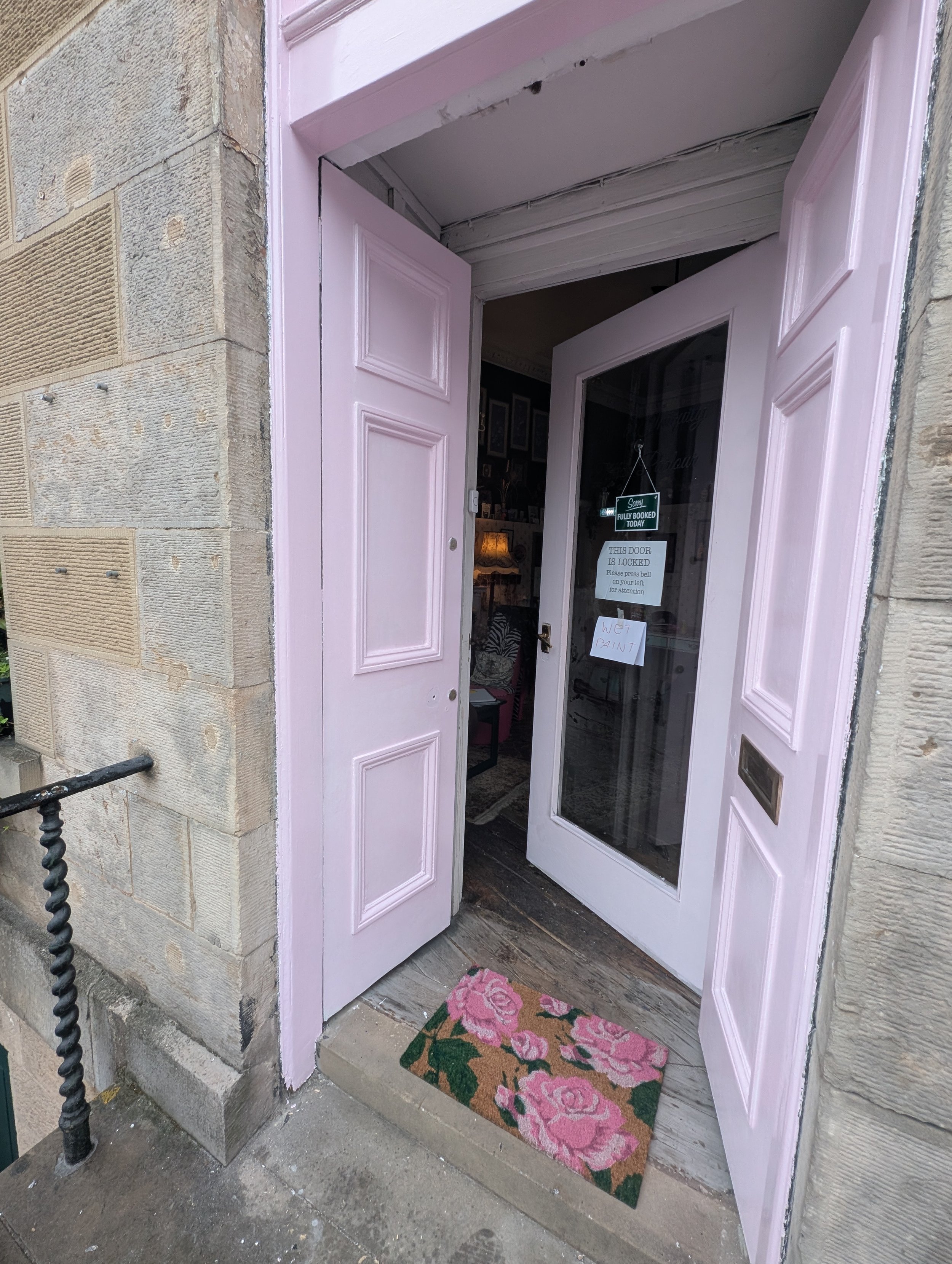 Open pink door with glass panel, welcome mat with pink roses, and signs indicating the door is locked and wet paint inside.