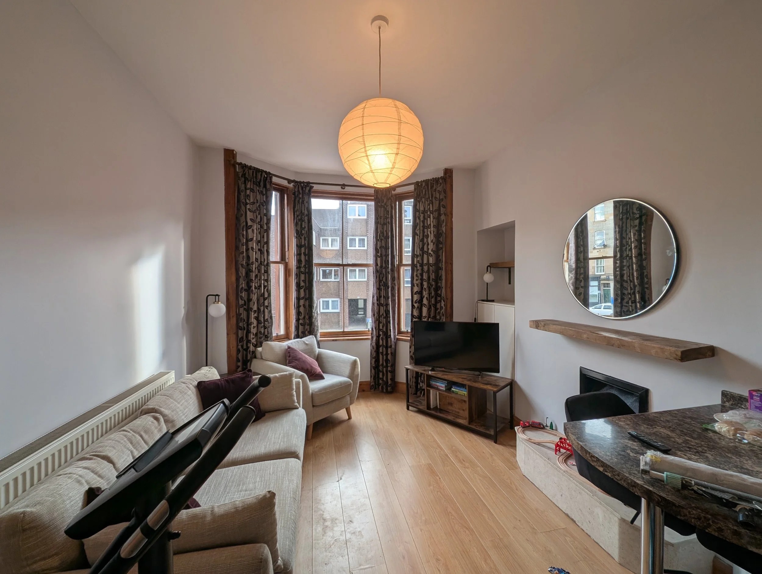 Living room with a large bay window, beige sofa, armchair with pillows, a TV on a wooden stand, a round mirror on the wall, a wooden mantel, and a dining table with chairs. There is a paper lantern ceiling fixture and a partial view of baby strollers