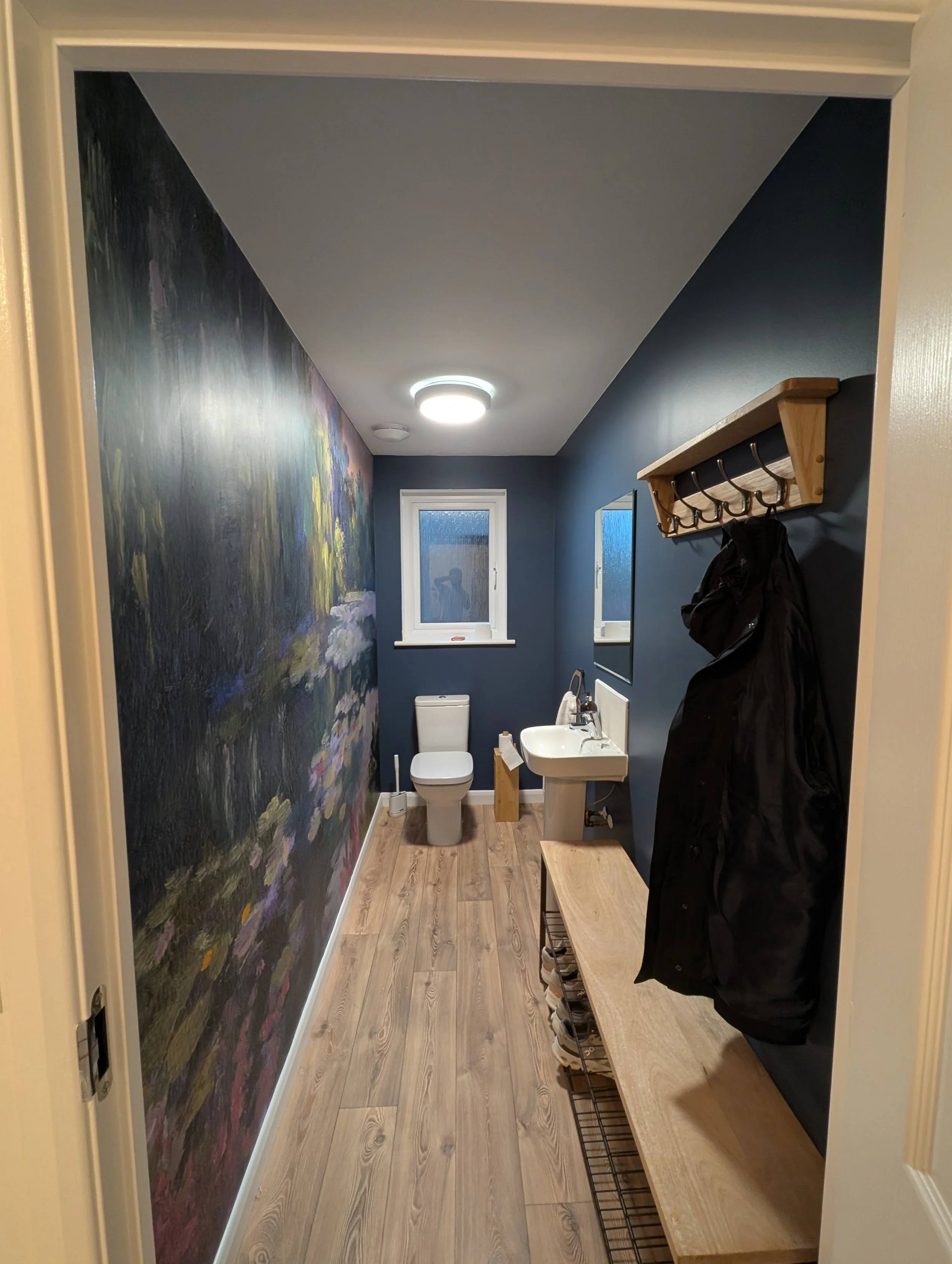 A small bathroom with blue walls, a toilet, a small white sink, a mirror, and a wooden bench. There is a water-resistant dark-colored coat hanging on a wooden hook rack on the wall to the right. The left wall features a large mural with floral and na