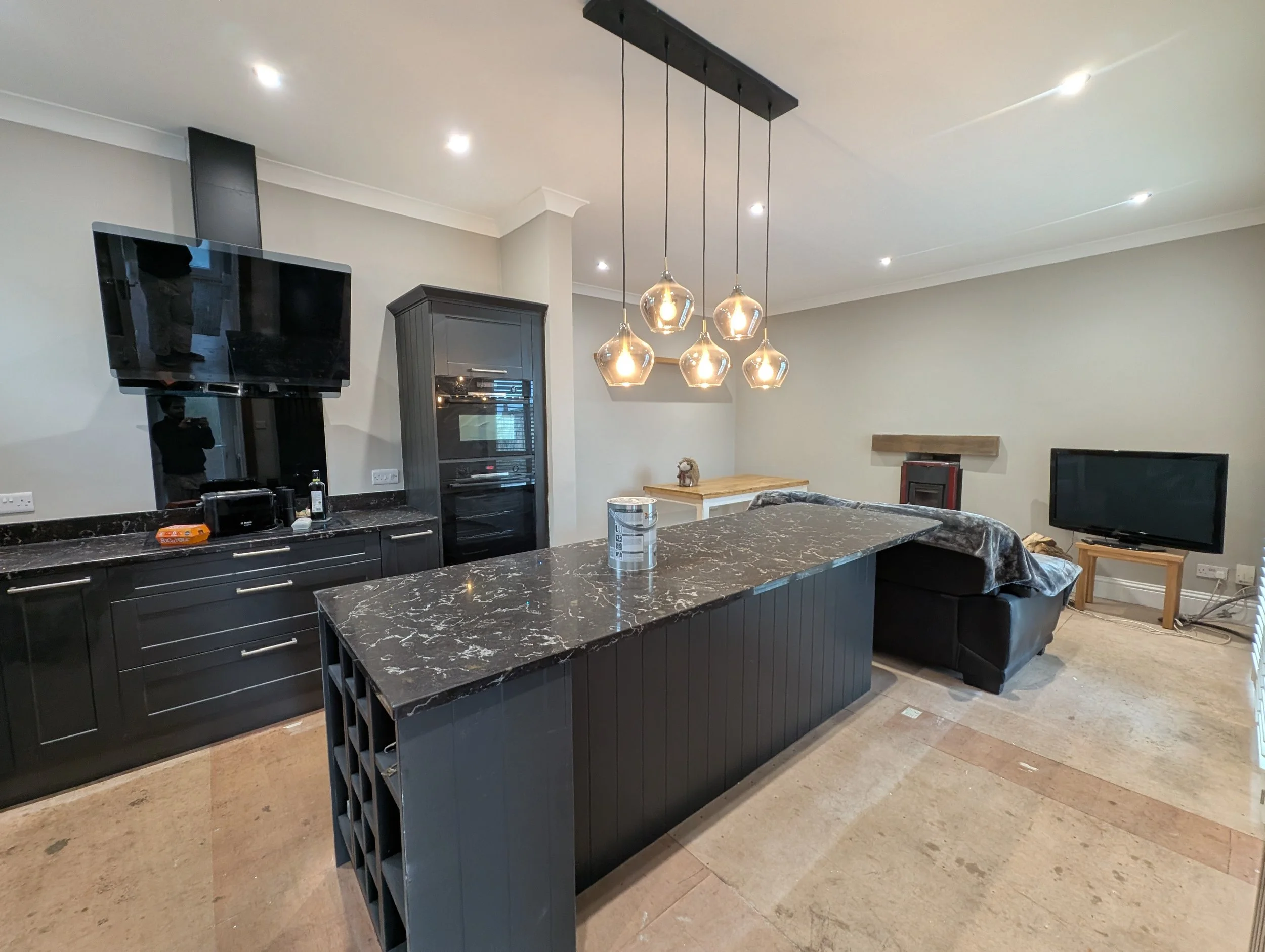 Living room with black kitchen cabinetry, a marble-topped island, a black sofa, a wall-mounted TV, and modern pendant lighting.