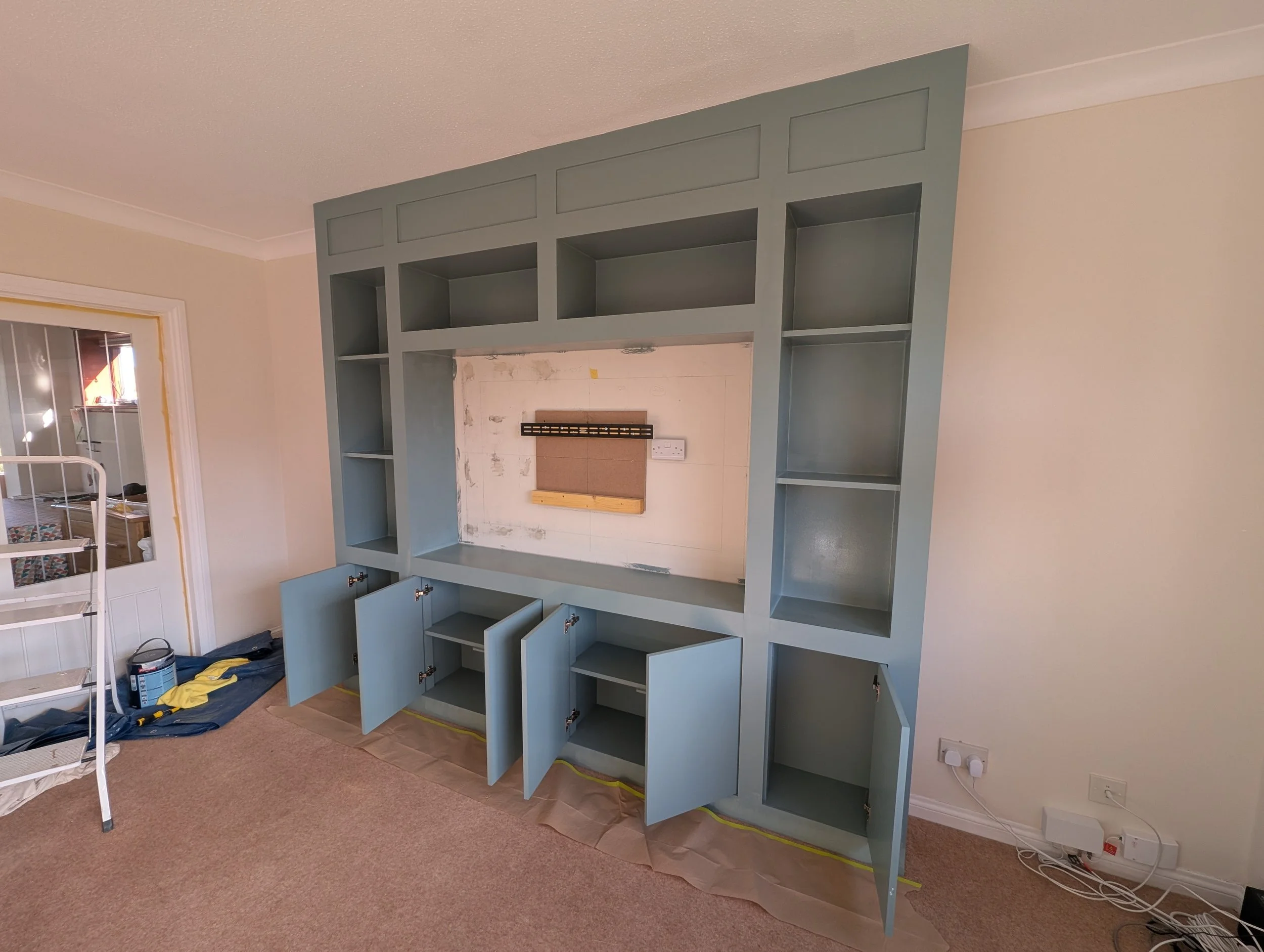 A partially assembled blue built-in entertainment center with open cabinets and shelves in a living room, with some cabinets open and painting supplies on the floor.