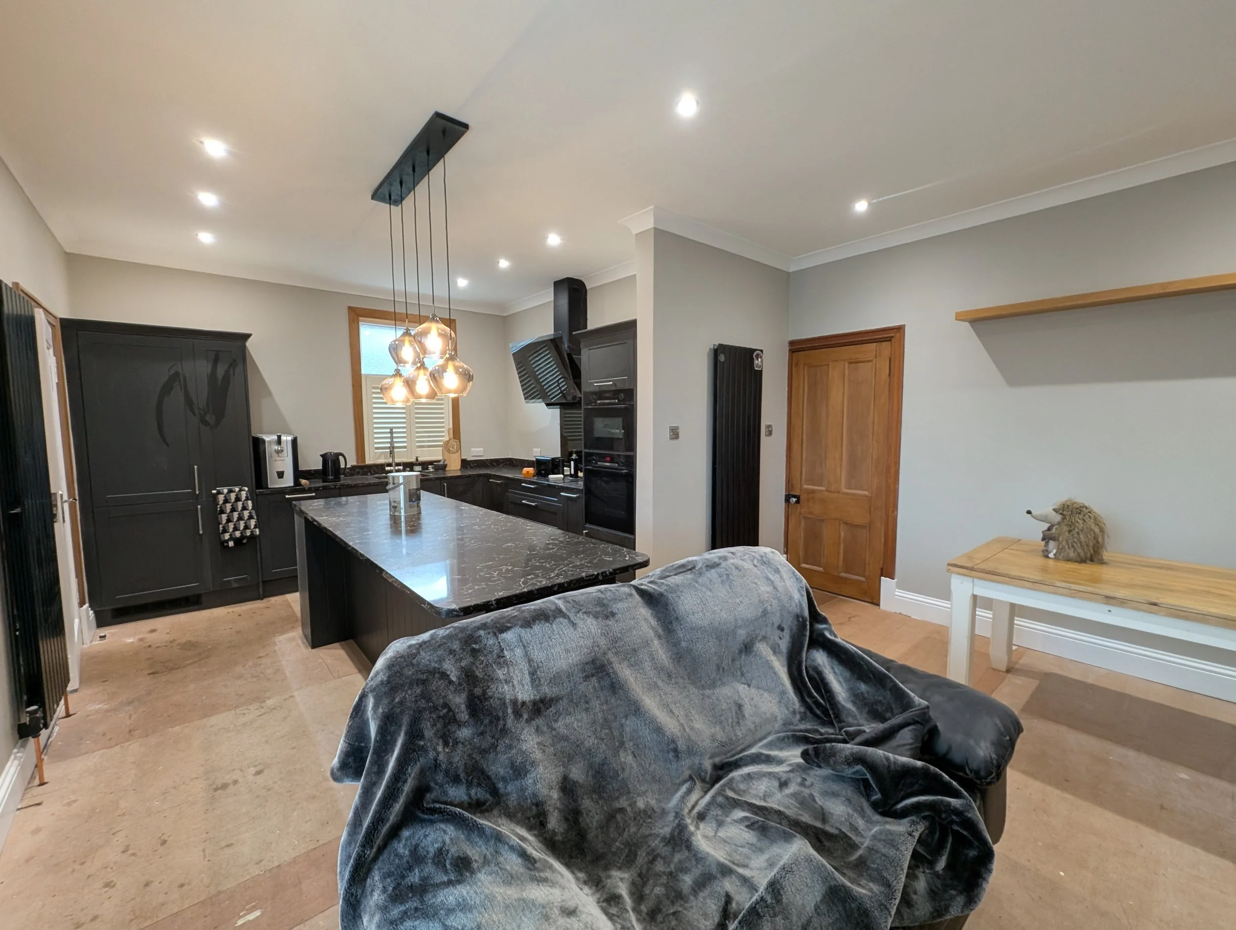 Open kitchen with black cabinets, marble island, and a sofa in the foreground. Pendant lights hang above the island. There is a wooden door, a small table with a hedgehog ornament, and a window with shutters.