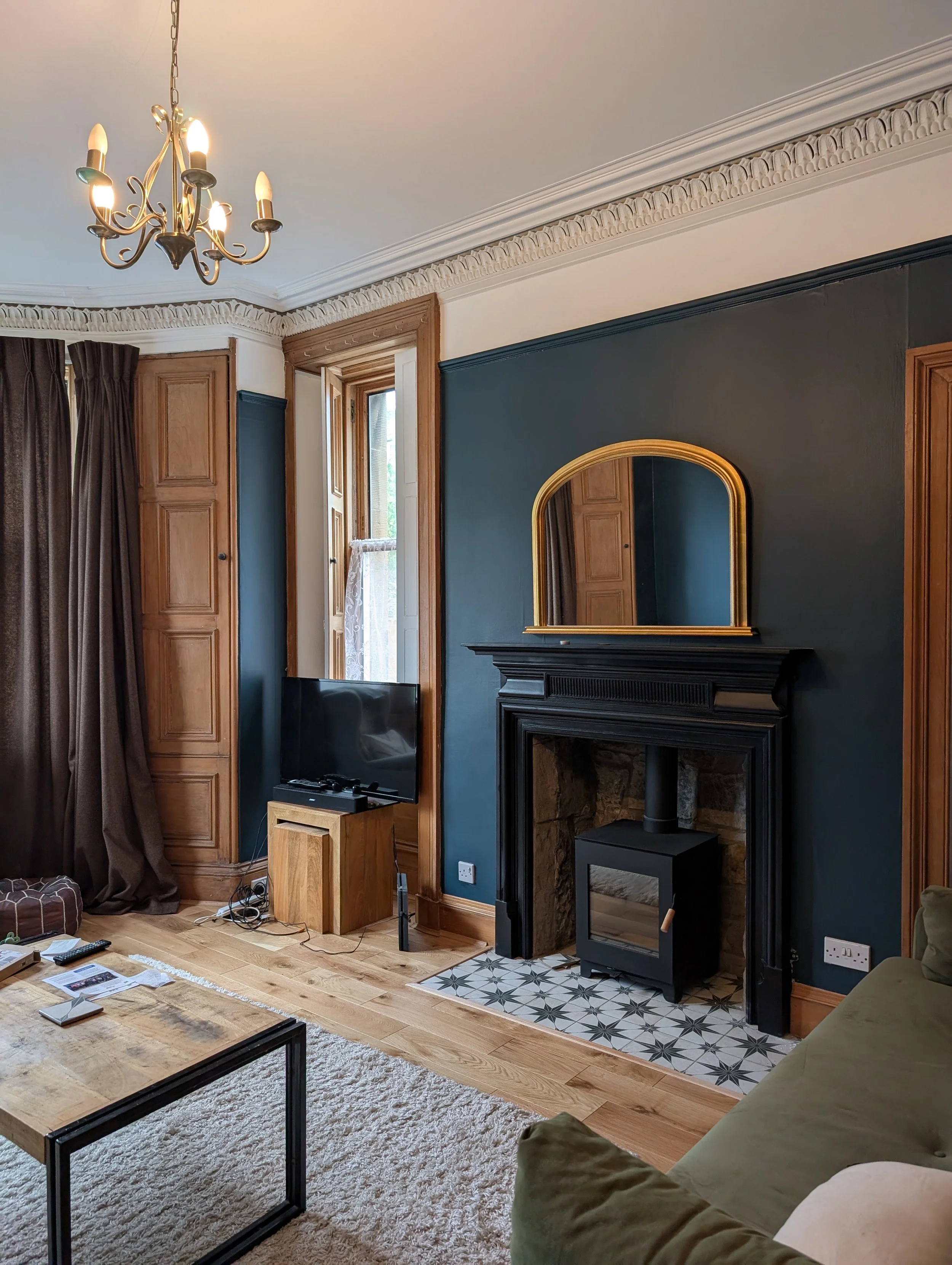 Living room with a black fireplace, blue wall, wooden window frame, TV on a wooden stand, green sofa, rug, and chandelier.