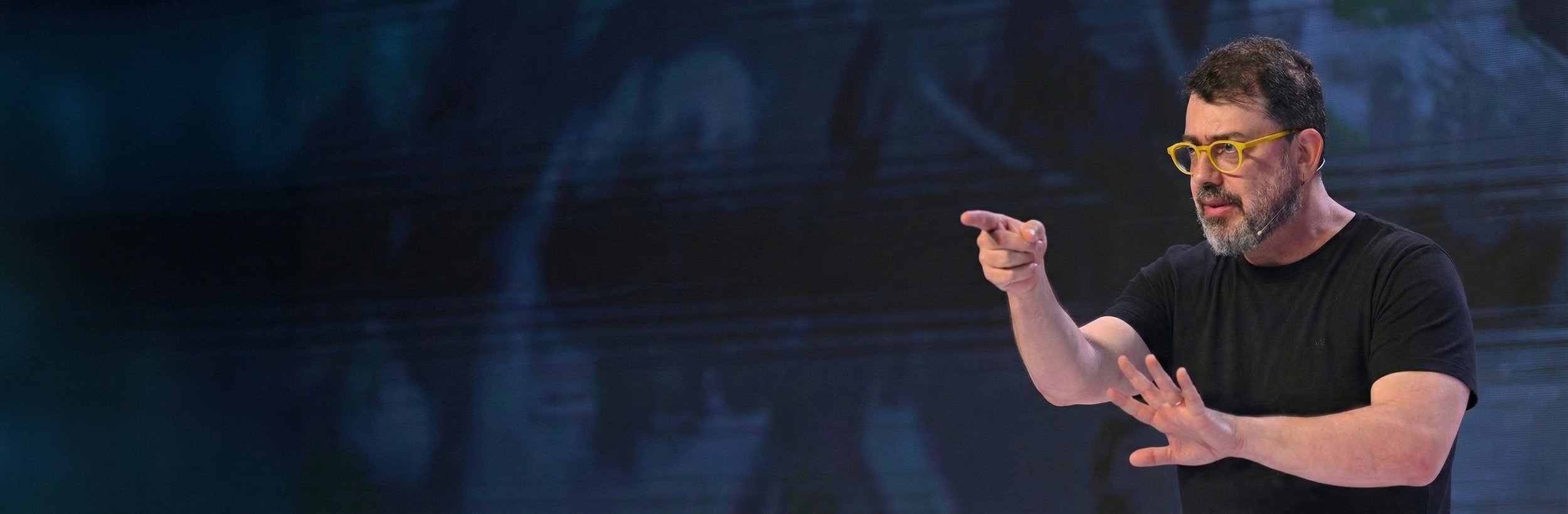 Fotografia de Pedro Doria, um homem com barba e óculos amarelos, vestindo camiseta preta, aponta com o dedo durante uma palestra, com no fundo uma grande tela de led e elementos de palco.