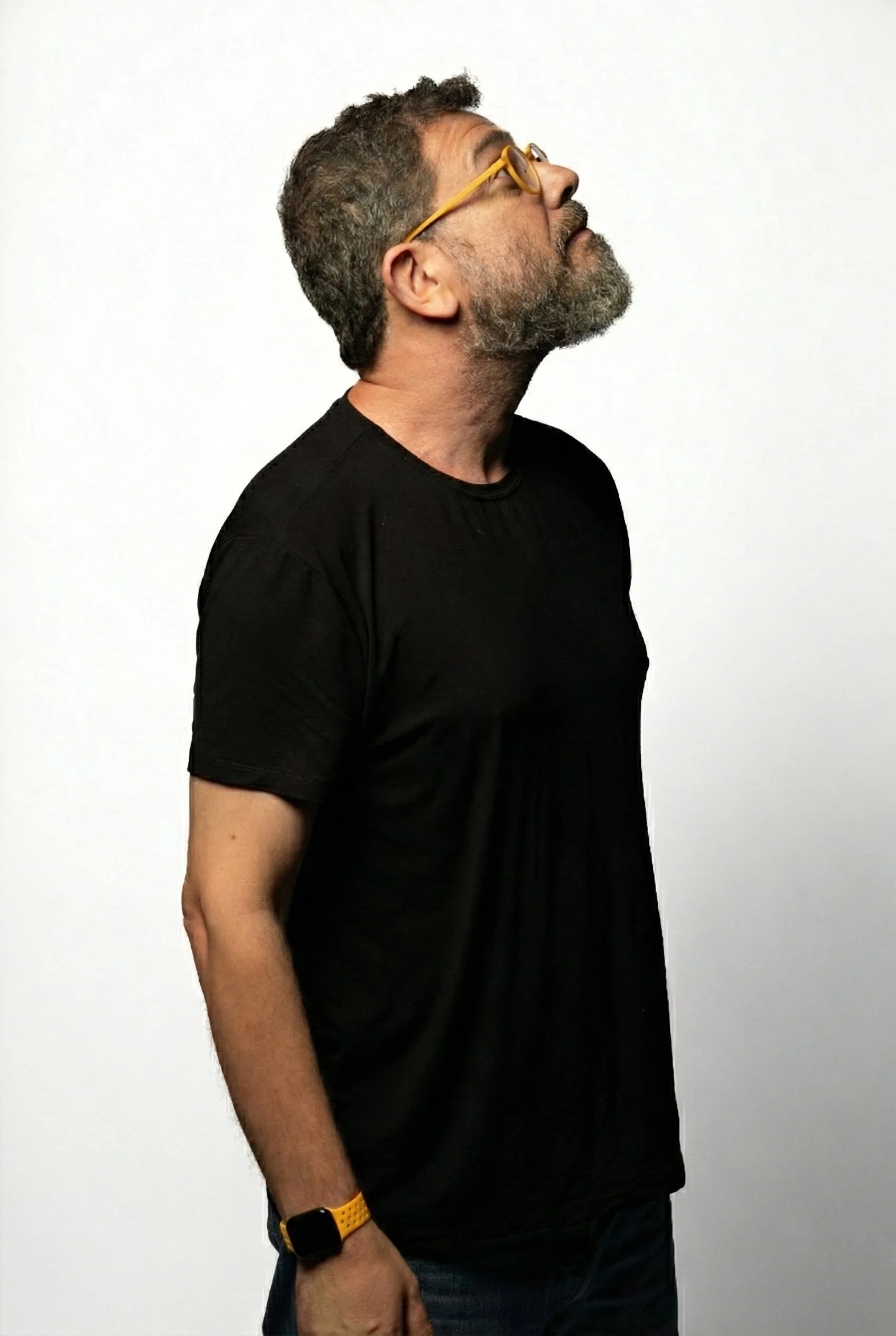 Pedro Doria, um homem de cabelo escuro, barba grisalha, usando óculos amarelos, camiseta preta, olhando para cima, com fundo branco.