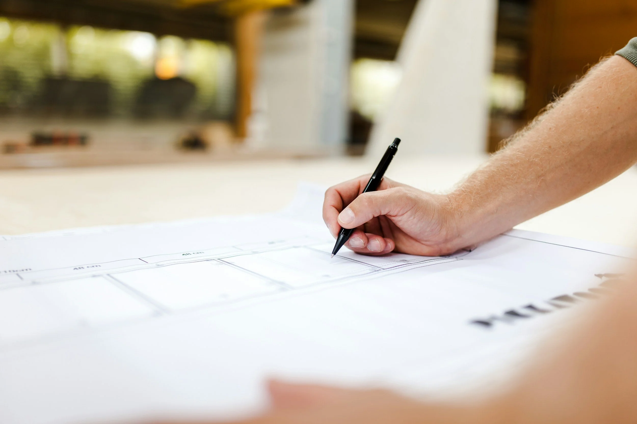 A person with light skin and freckles is using a black pen to write or draw on a large sheet of paper with blueprints or architectural plans printed on it. The background is blurred, showing an outdoor setting with a wooden structure and greenery.