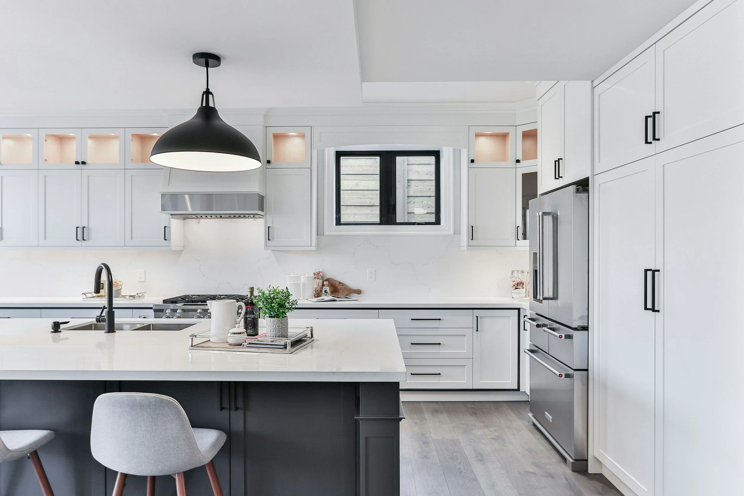 Modern white kitchen with black hardware, stainless steel appliances, a central island with a white countertop, black pendant light, and a window above the sink