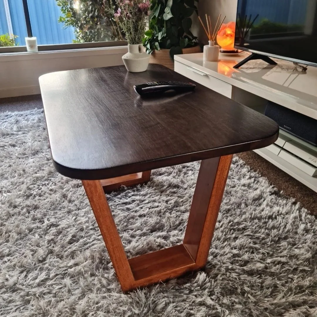 A modern living room with a dark wooden table, decorated with a white flower vase, a TV remote, and a diffuser. The room features a fluffy gray rug, a white console with a salt lamp and a flat-screen TV, and a large window with greenery outside.