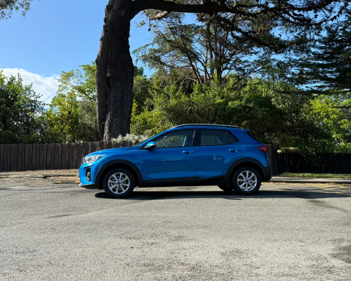 A blue compact SUV parked on a paved street, with a wooden fence and large trees with green foliage in the background under a partly cloudy sky.