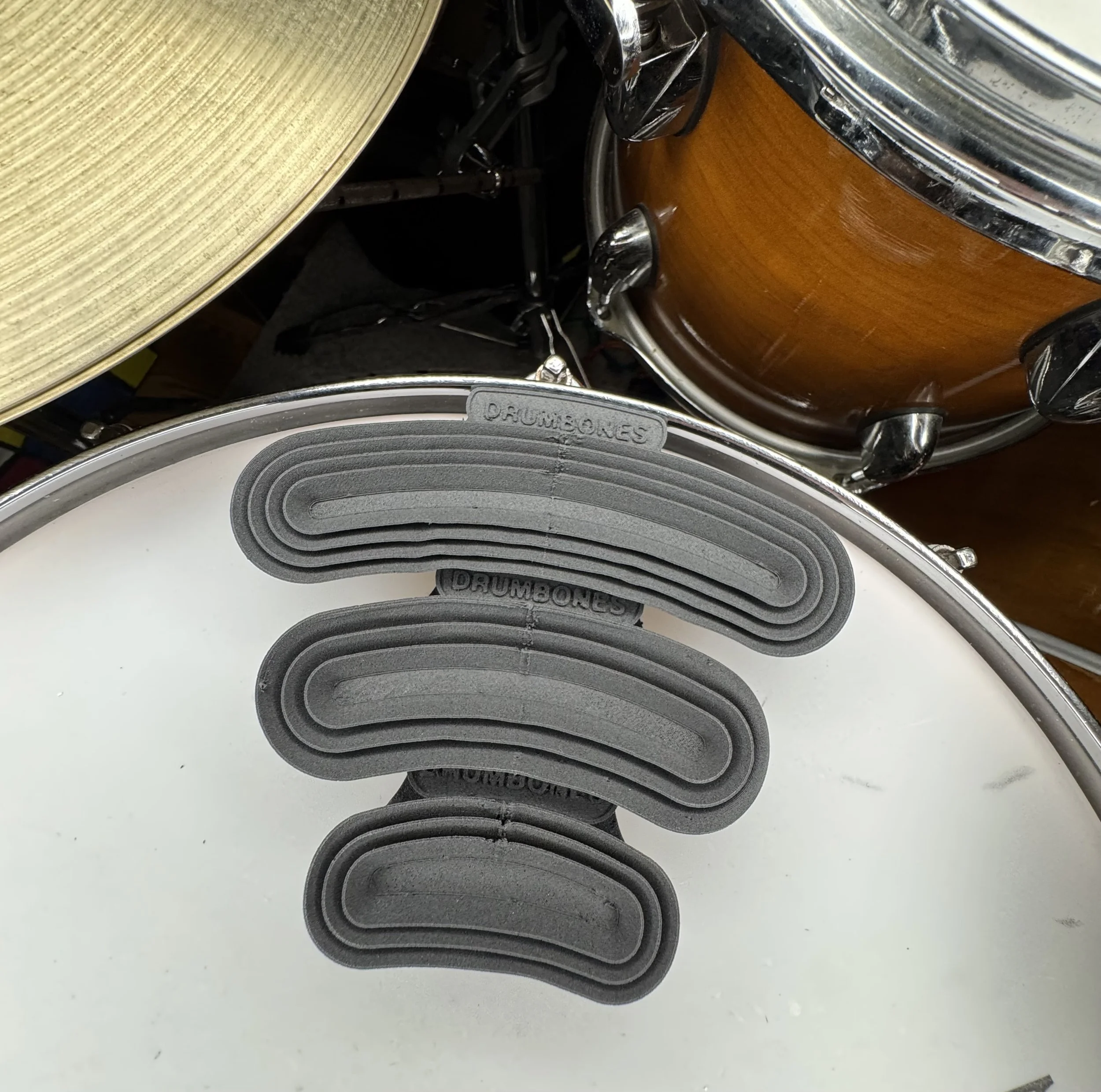 Close-up of a bass drum with a white drumhead and grey foam pads labeled 'DRUMONES' attached to the drum rim, with parts of a cymbal and a wooden tom drum visible in the background.