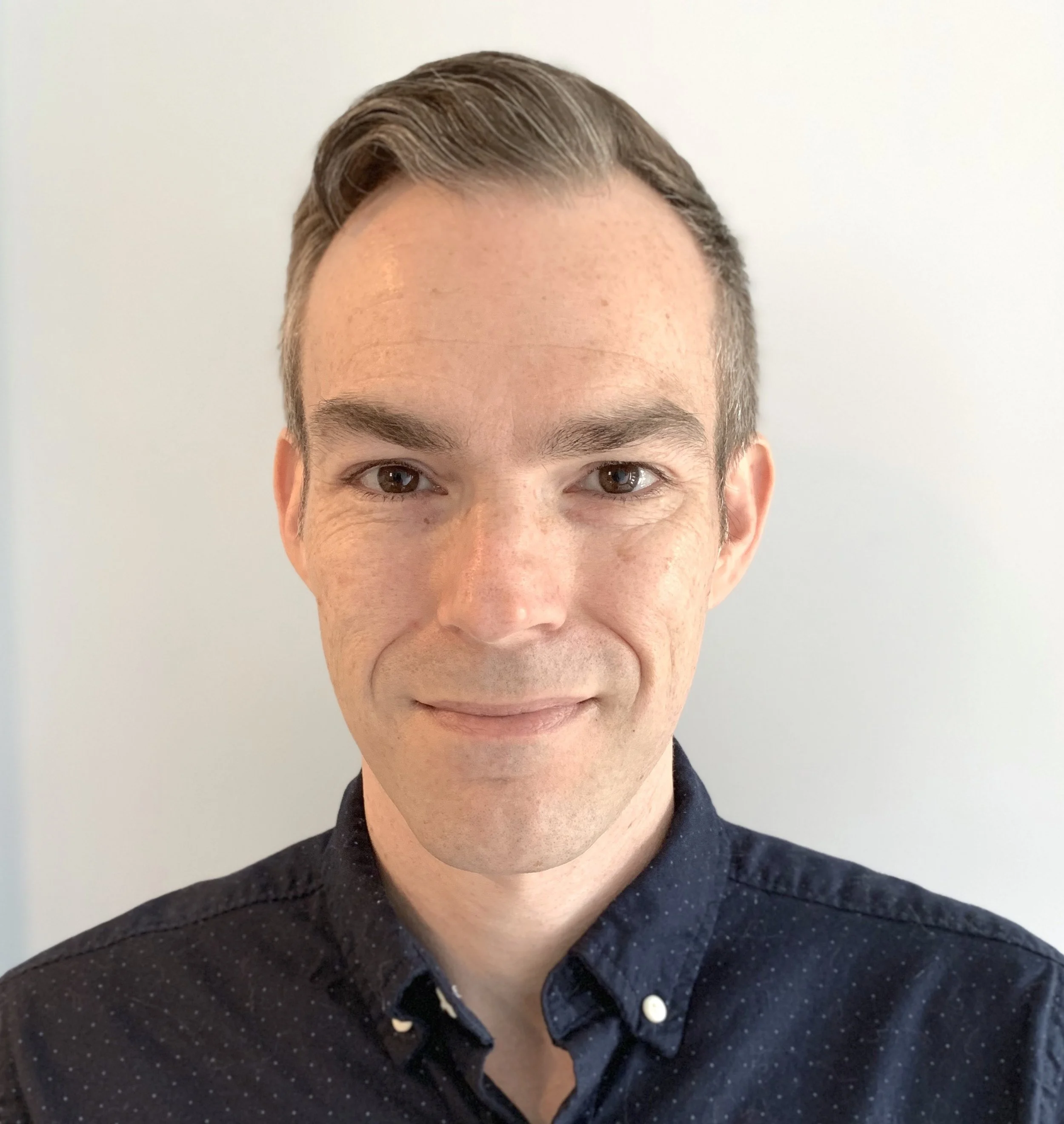 Headshot of a man with short, well-groomed hair, wearing a navy blue button-up shirt, smiling softly against a plain white background.