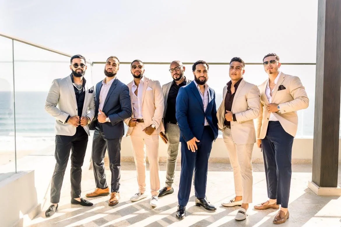 Group of eight men dressed in suits standing outdoors on a balcony with a view of the ocean.