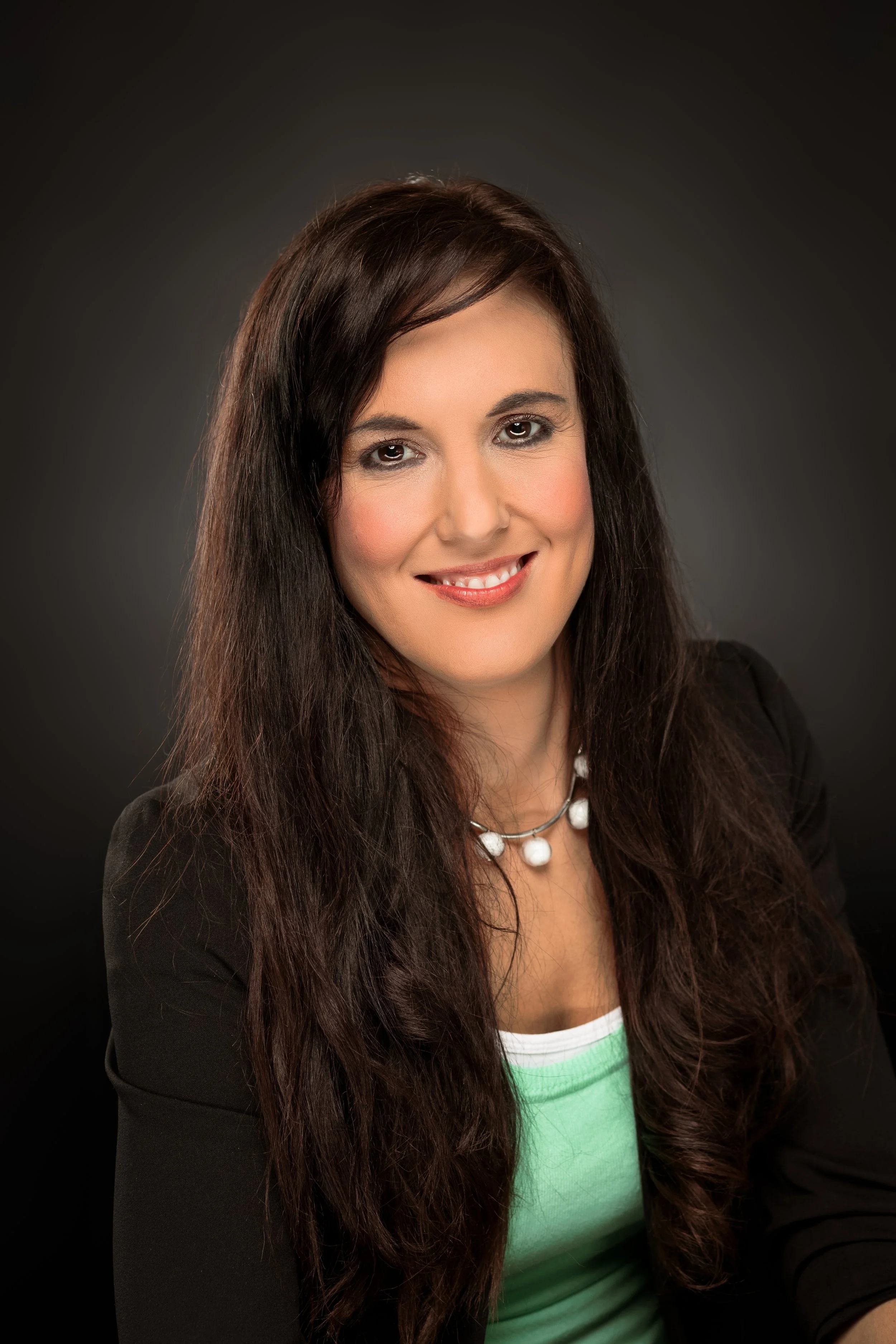 A woman with long dark hair smiling, wearing a black blazer, a green top, and a white necklace against a dark background.