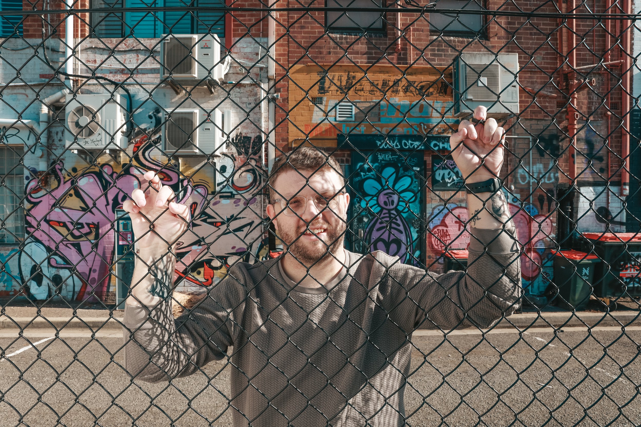 A man with glasses and tattoos on his arms smiling and holding onto a chain-link fence, with graffiti art on brick buildings in the background.