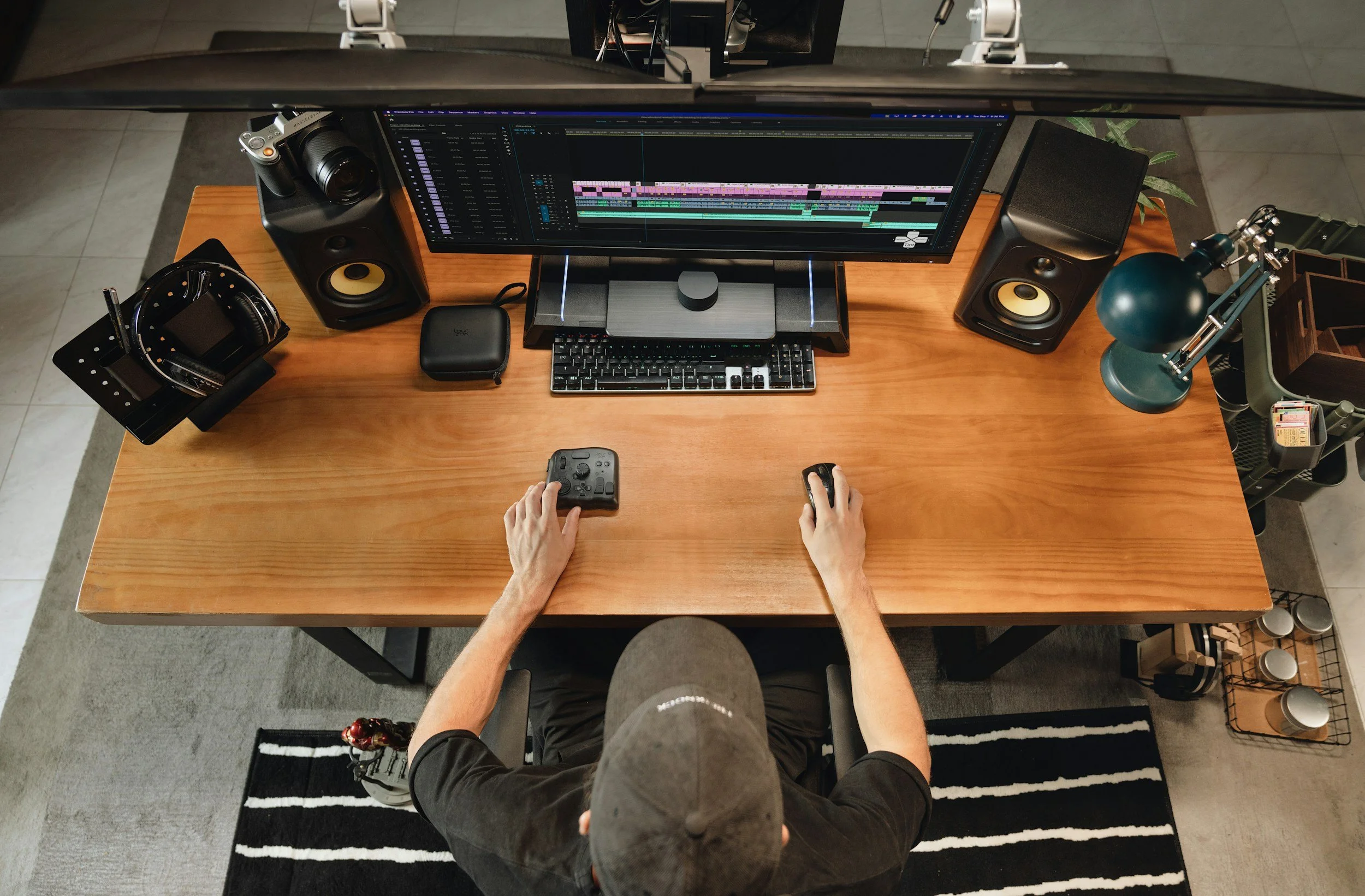 Overhead view of a person working at a wooden desk with a large monitor displaying video editing software, surrounded by studio speakers, a camera, headphones, a lamp, and other work accessories.