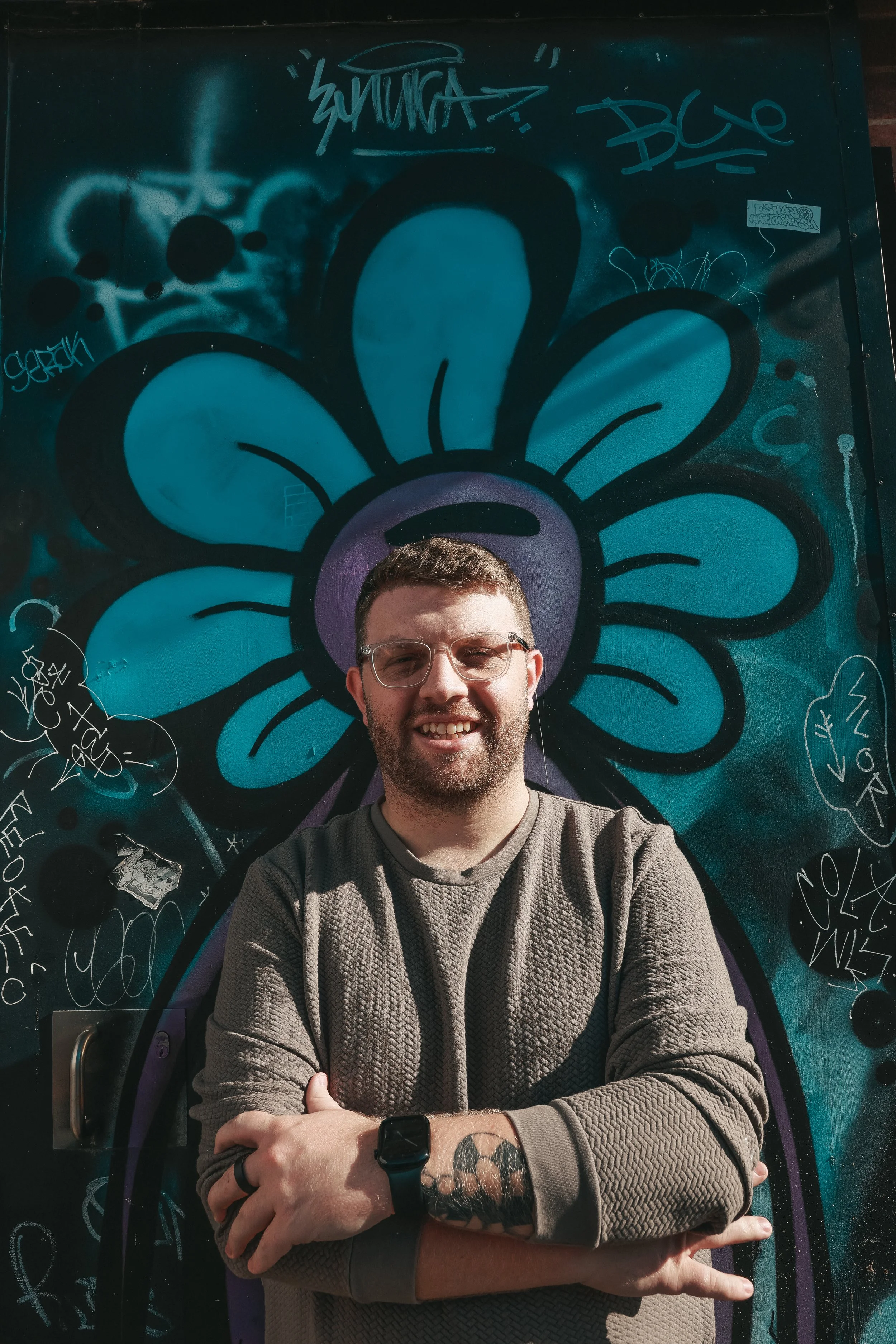 A young man with glasses and a beard smiling, standing in front of a colorful graffiti mural of a blue flower on a dark wall, with arms crossed and a tattoo on his left forearm.