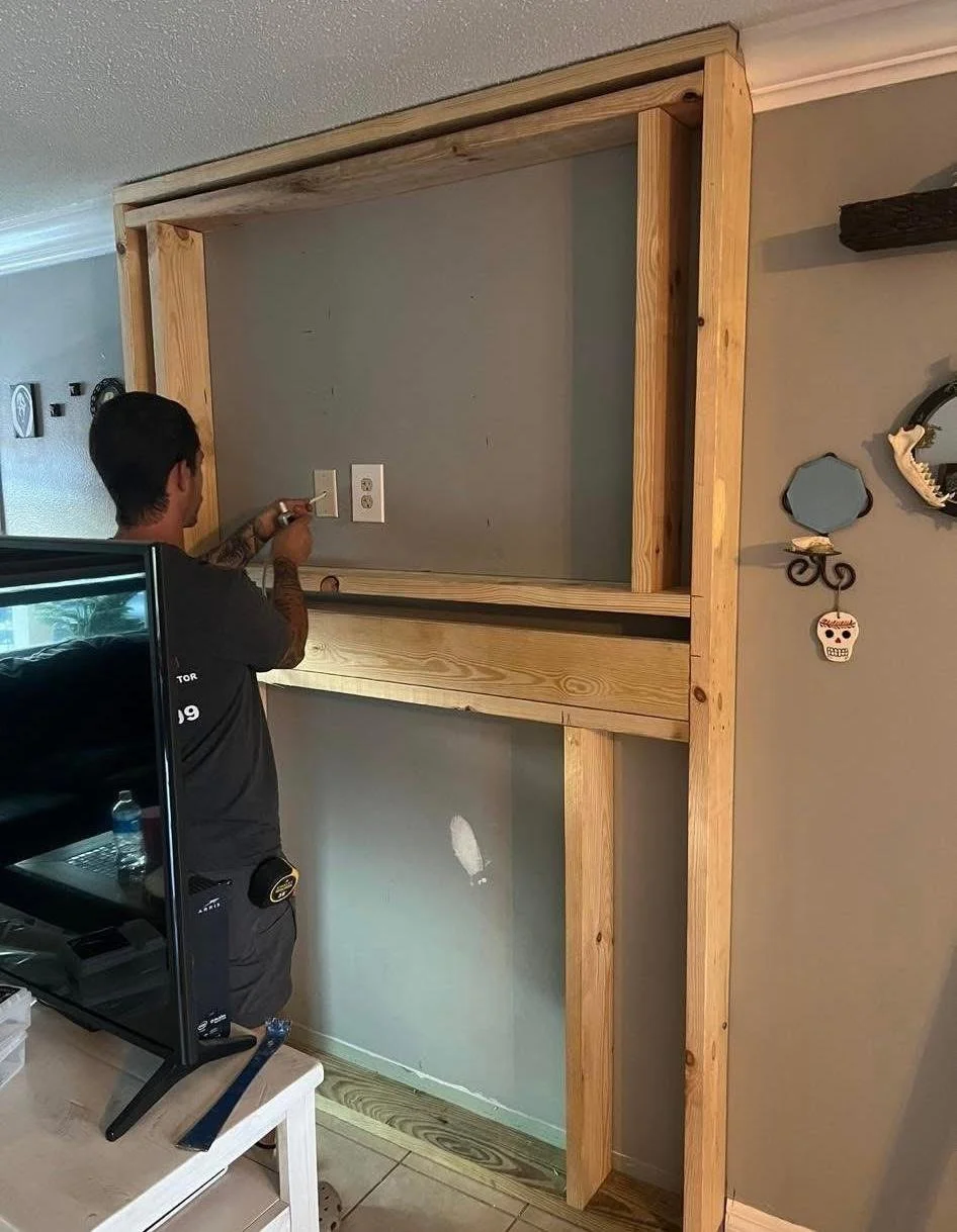 A person working on a wooden framework for a wall or cabinet in a room, with a TV and some wall decor visible.