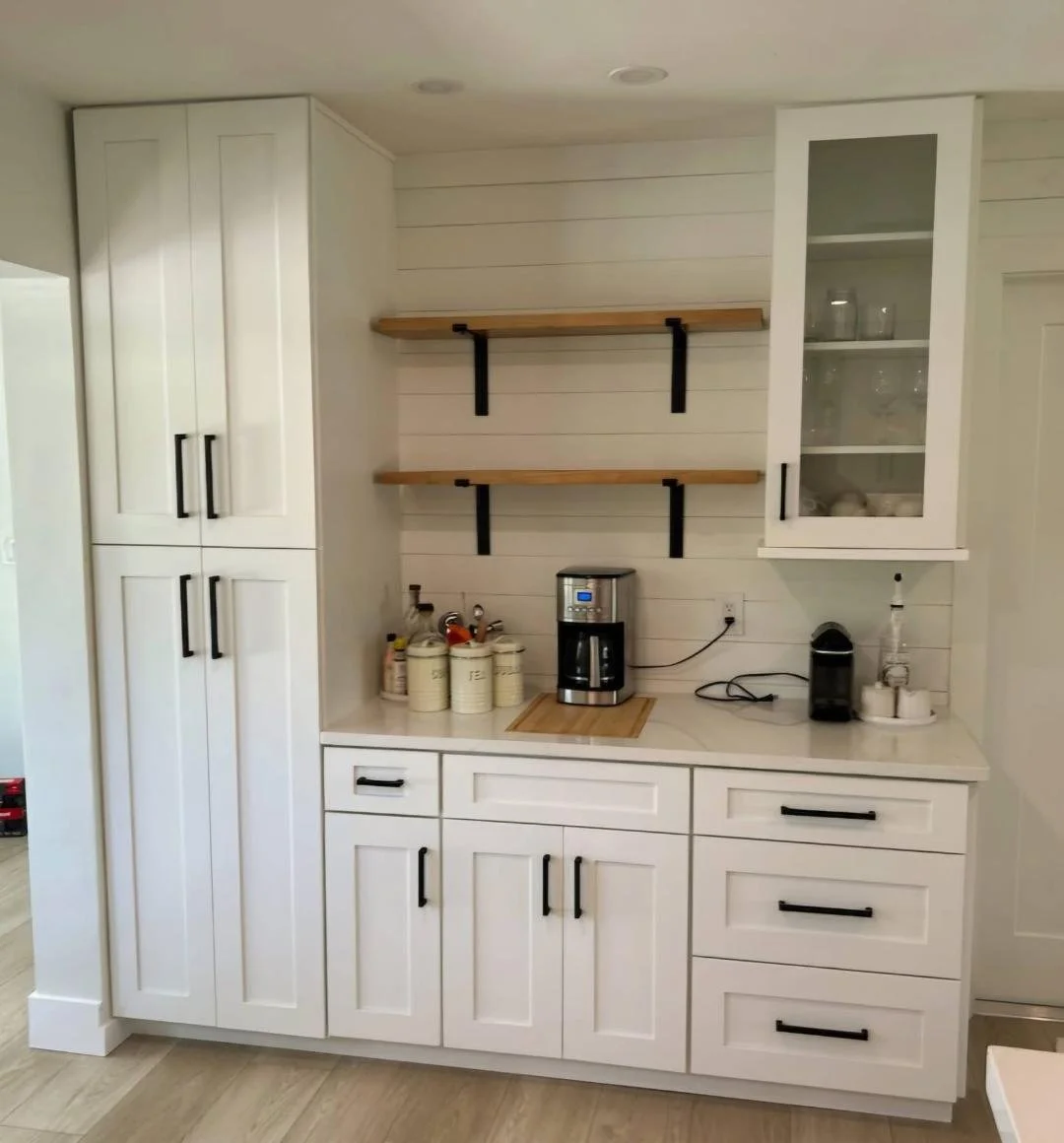 White kitchen cabinet with black handles, open shelves, countertop with coffee maker, utensils, and storage jars, white shiplap wall, and wooden shelves with black brackets.