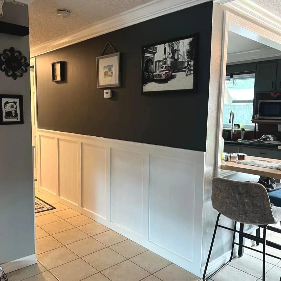 Interior of a home showing a dark accent wall decorated with framed artwork and a view into the kitchen with a window and kitchen appliances.