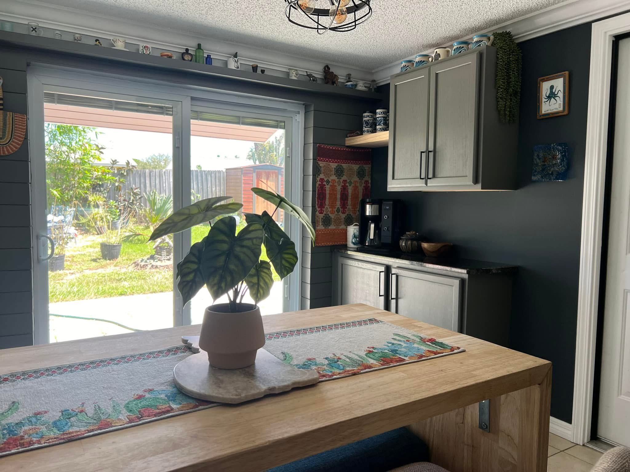 A kitchen dining area with a wooden table, a potted plant with large green leaves, a sliding glass door opening to a backyard with plants, a black coffee machine, wall-mounted cabinets, and decorative items on the shelves and ceiling.