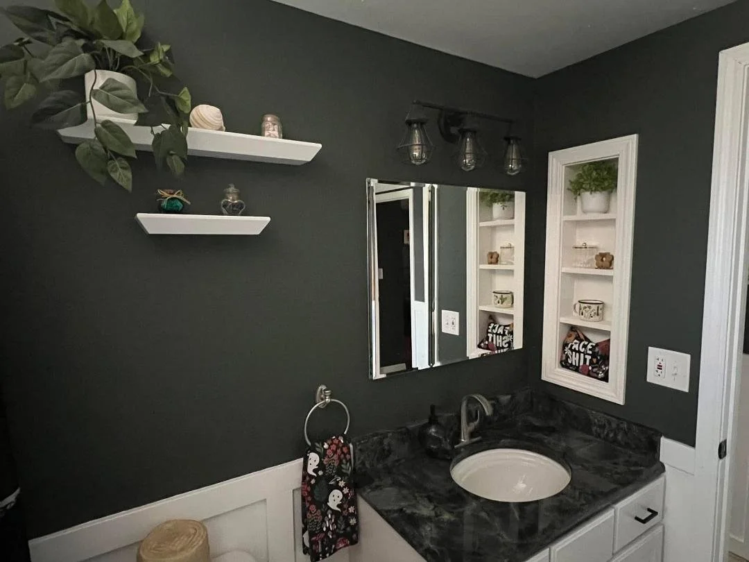 Bathroom with dark gray wall, white shelves with decorative items, a mirror, a black marble countertop with a beige sink, and a small shelf with a green plant.