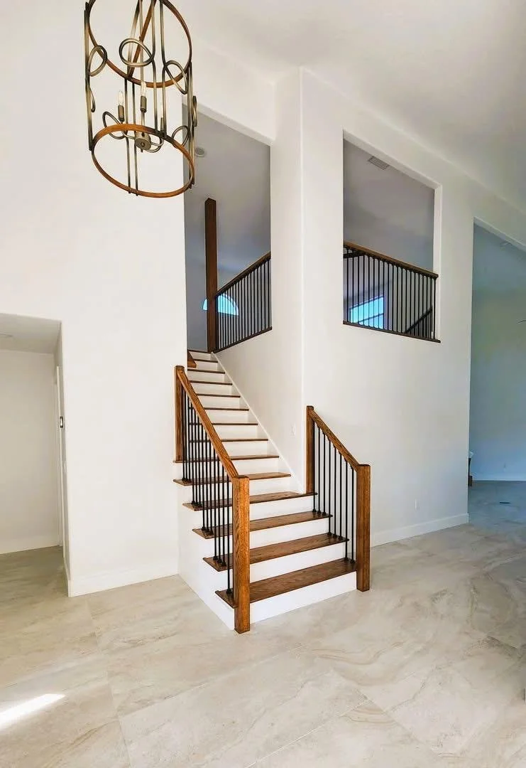 Interior of a modern home with a staircase featuring wooden handrails and black metal balusters, beige tile flooring, and a white wall. A decorative chandelier hangs from the ceiling.