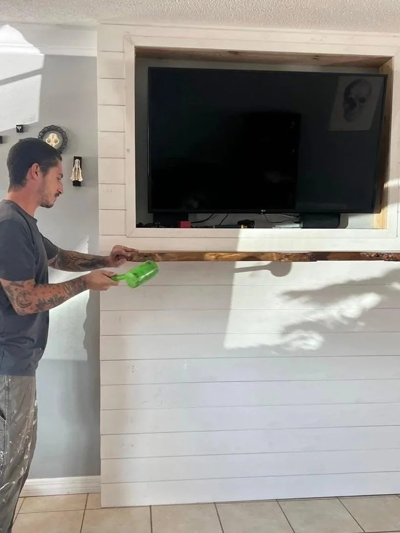 A man with tattoos on his arms is holding a green spray bottle and spraying a wooden shelf in front of a wall-mounted flat-screen TV in a room with light-colored walls and tiled floor.