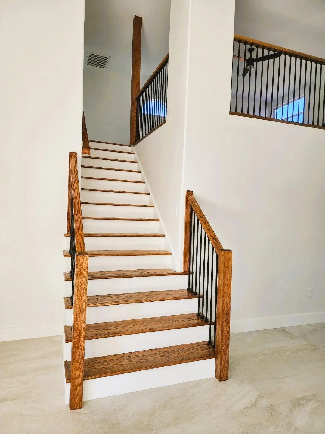 Interior view of a staircase with wooden steps, black metal balusters, and a wooden handrail, leading to an upper floor with a balcony.