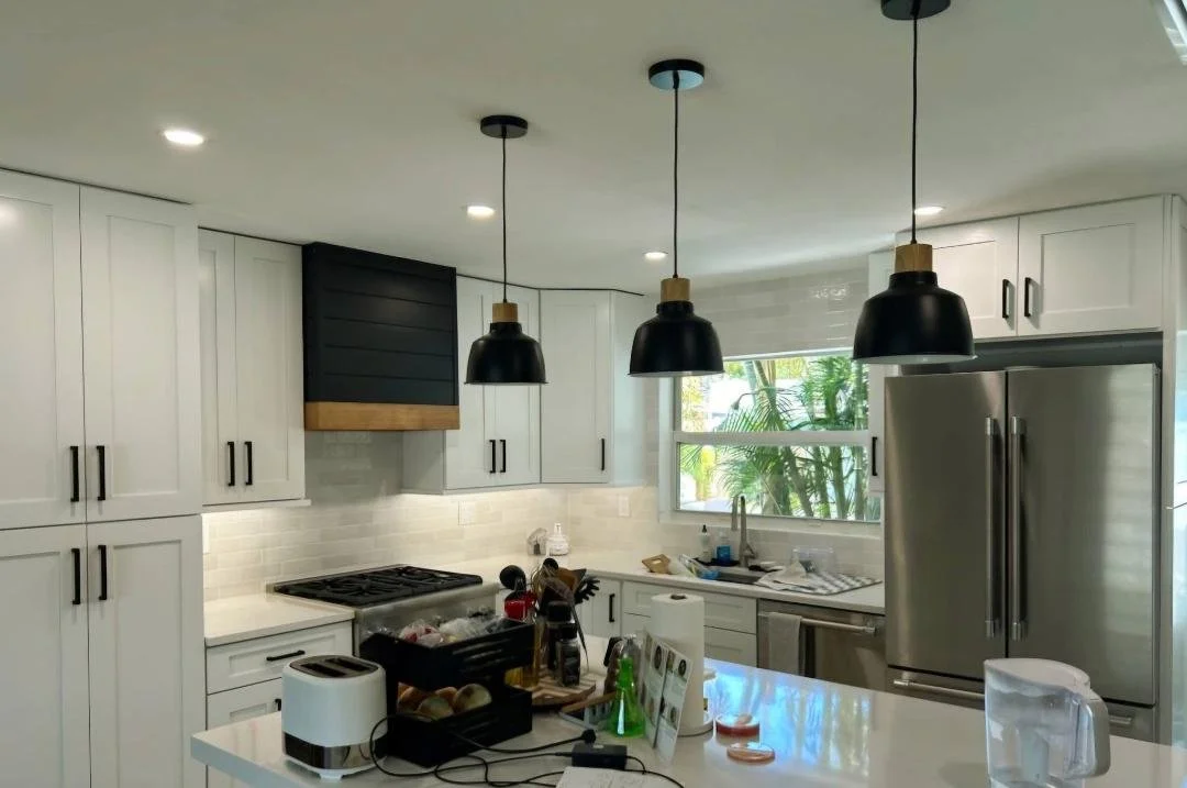 Modern kitchen with white cabinets, stainless steel refrigerator, black pendant lights, and a window overlooking greenery.
