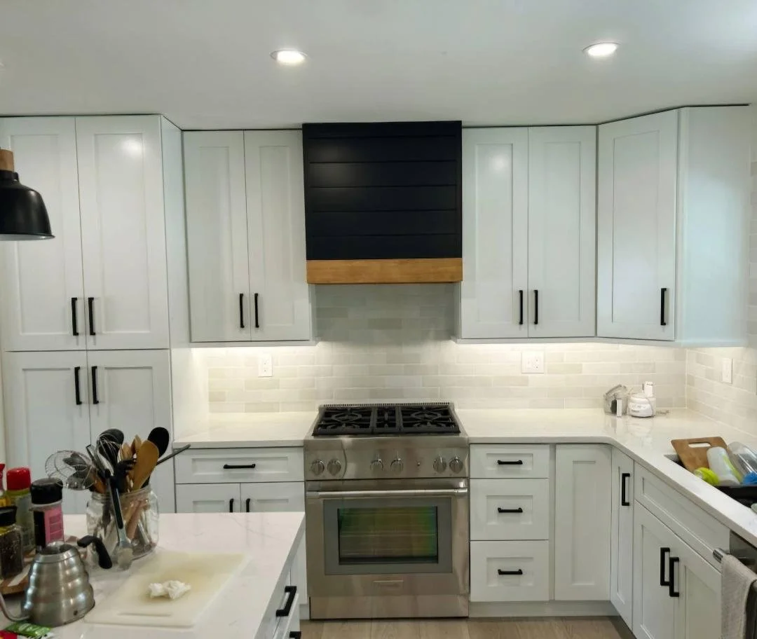 Modern kitchen with white cabinets, black handles, grey backsplash, and stainless steel oven; black range hood with wood trim; countertop clutter including utensils, a kettle, and cleaning supplies.