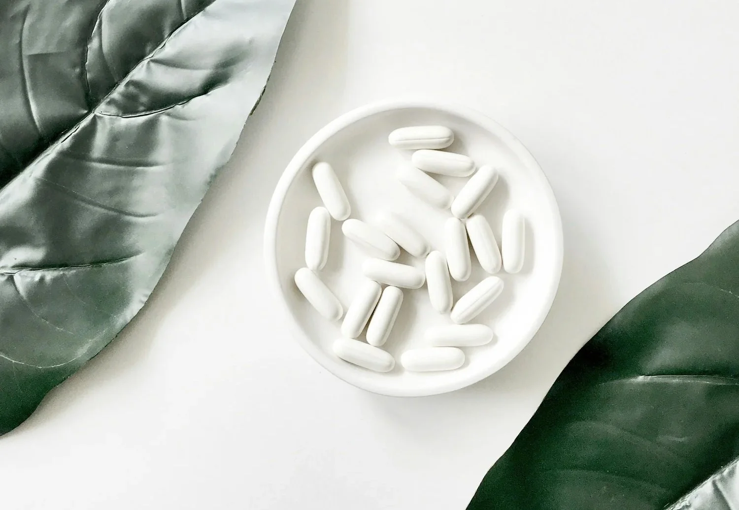 White capsules in a round white dish placed on a white surface, flanked by green and silver leaves.