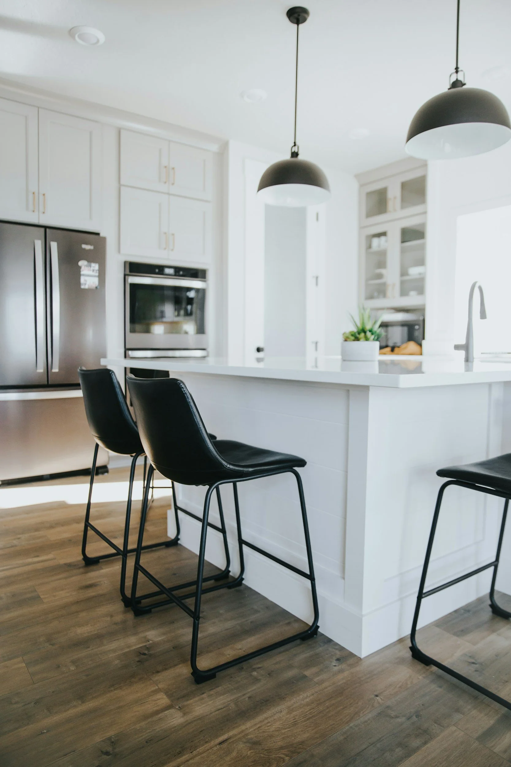 Modern white kitchen with black bar stools at a white island, stainless steel appliances, pendant lighting, and wood flooring.